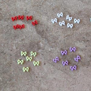 May include: A collection of small, colorful bow-shaped embellishments. The bows are in various colors, including blue, red, white, pink, green, purple, and light blue. They are arranged in small groups on a textured, neutral-toned surface.