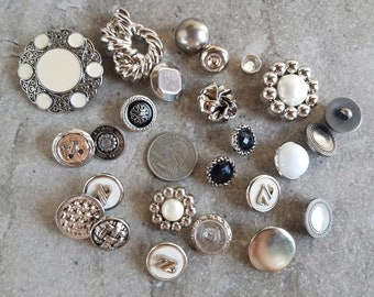 May include: A collection of various vintage buttons in silver, black, white, and pearl. The buttons feature different designs, including floral, geometric, and textured patterns. Some buttons have a pearl or black gem center. A quarter is included for size reference.