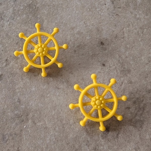 May include: A pair of bright yellow ship wheel shaped objects. Each piece features a central hub with eight spokes radiating outward, ending in small rounded knobs. The design mimics a nautical steering wheel with a smooth, matte finish.