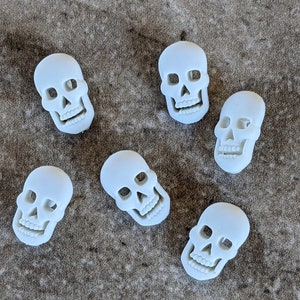 May include: Six white ceramic skull-shaped ornaments. The skulls have a smooth, matte finish and are all facing forward.