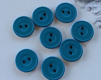 May include: A collection of eight teal-colored, round buttons with two holes each. The buttons are arranged on a light gray and white speckled surface. These buttons are suitable for sewing and craft projects.