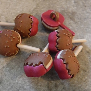 May include: Six brown and white candy buttons with red frosting and a white stick. The buttons are shaped like a candy apple.