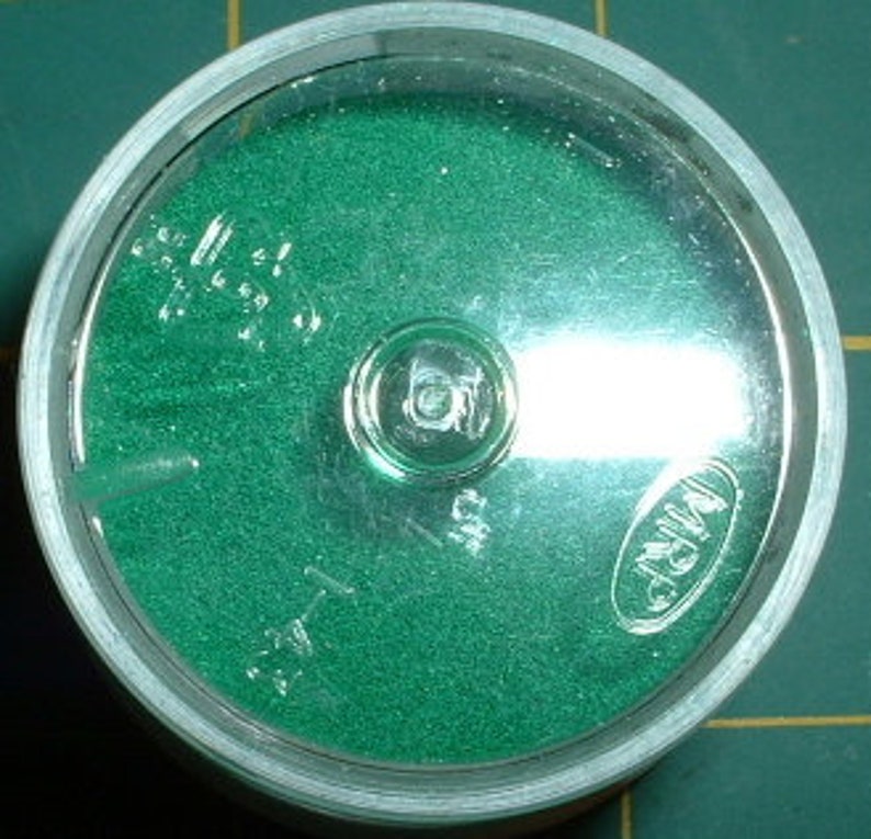 May include: A small, round, clear plastic container with a green, textured surface inside. The container has a clear lid and a small, silver button in the center. The text "WINDSOR" is printed on the lid.