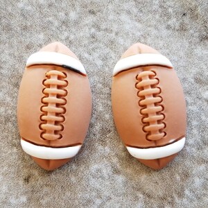 May include: Two brown and white football-shaped ornaments with a white stitched seam down the center.