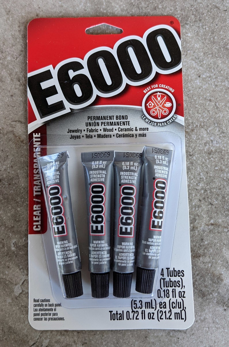 May include: A retail package containing four 0.18 fl oz tubes of E6000 industrial strength adhesive. The red and white card features the E6000 logo, lists applications like jewelry, fabric, wood, and ceramic, and states a total of 0.72 fl oz.