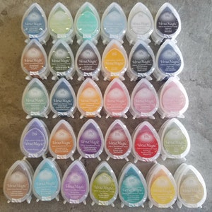 May include: A collection of 27 ink pads in various colors, each with the text "VersaMagic" and "Color Name" printed on the front. The ink pads are shaped like teardrops and are arranged in rows on a gray surface.