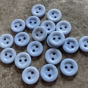 May include: A collection of light blue plastic buttons with two holes and a decorative design. The buttons are arranged in a random pattern on a textured surface.