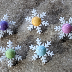 May include: Five white snowflake ornaments with glitter centers. The centers are purple, yellow, pink, green, and blue.