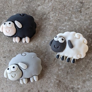 May include: Three small, handmade sheep figurines. One is black, one is gray, and one is white. They are all made of a soft, pliable material and have cute, cartoon-like faces.