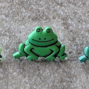 May include: Three green frog buttons with black eyes and a black smile. The frogs are sitting on a light gray surface.