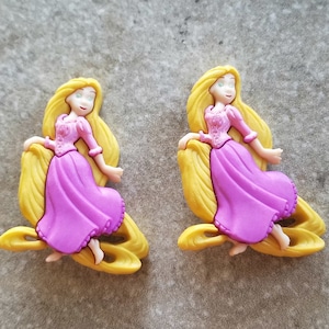 May include: Two plastic figurines of Rapunzel from the Disney movie Tangled. The figurines are in a standing pose with long blonde hair and are wearing purple dresses.