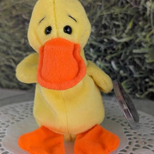 May include: A yellow plush duck with orange feet and beak. The duck has large black eyes and a sad expression. The duck is sitting on a white doily.