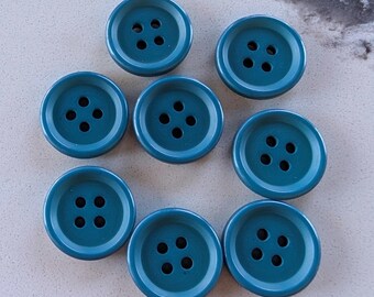 May include: A collection of eight teal-colored buttons. Each button is round with four holes in the center and a slightly raised outer rim. The buttons are arranged in a circular pattern on a light-colored surface.