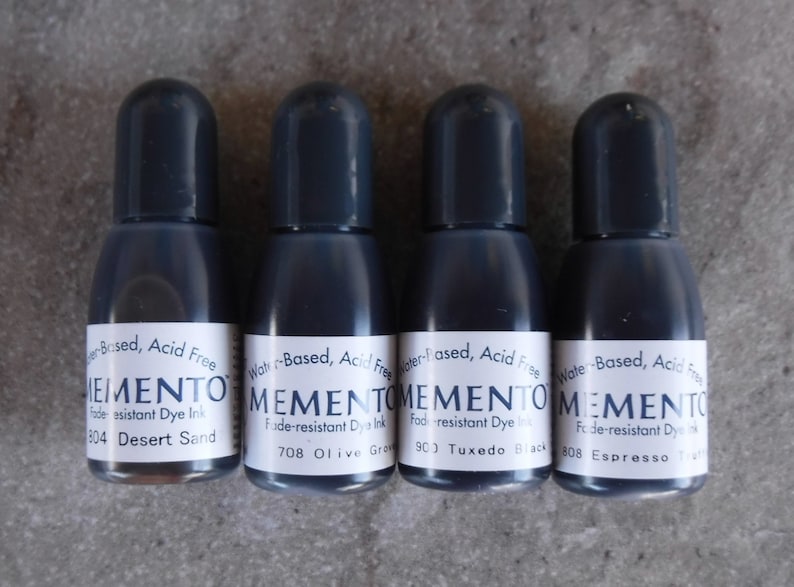 Puede incluir: Cuatro peque&ntilde;os frascos de tinta gris oscuro con tapas negras est&aacute;n alineados. Cada frasco tiene una etiqueta blanca con la palabra "MEMENTO" y el nombre del color de la tinta. Las etiquetas tambi&eacute;n indican que la tinta es a base de agua, sin &aacute;cido y resistente a la decoloraci&oacute;n.