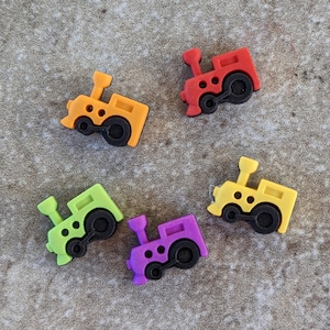 May include: Five colorful plastic toy train buttons in various colors, including red, orange, yellow, green, and purple. The buttons are shaped like trains with black wheels and details.