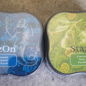 May include: Two ink pads with a decorative design. The ink pad on the left is blue and says "StazOn Blue Hawaii Bleu Hawai Hawai Azul". The ink pad on the right is green and says "StazOn Cactus Green Vert cactus Verde cactus".