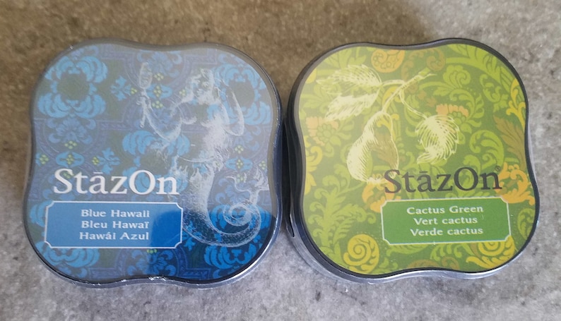 May include: Two ink pads with a decorative design. The ink pad on the left is blue and says "StazOn Blue Hawaii Bleu Hawai Hawai Azul". The ink pad on the right is green and says "StazOn Cactus Green Vert cactus Verde cactus".