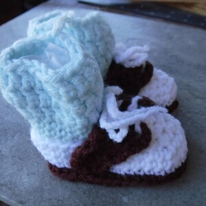 May include: A pair of crocheted baby booties with brown and white accents. The booties have a light blue cuff and white laces.