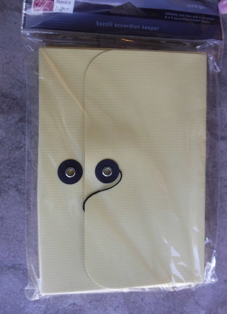 May include: A yellow accordion keeper with two black eyelets and a black string. The keeper is made of paper and has the text "bazill accordion keeper" printed on the front.