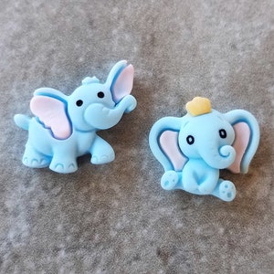 May include: Two blue elephant figurines with pink ears. One elephant is wearing a yellow crown.