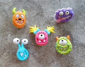 May include: Five colorful monster-themed buttons with different expressions. The buttons are made of plastic and have a flat back. They are perfect for adding a fun and whimsical touch to any craft project.