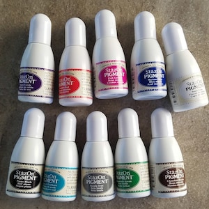 May include: A collection of ten small bottles of StazOn Pigment ink in various colors, including pink, red, blue, black, green, and brown. Each bottle has a white cap and a label with the brand name, ink color, and a description in English and Japanese.