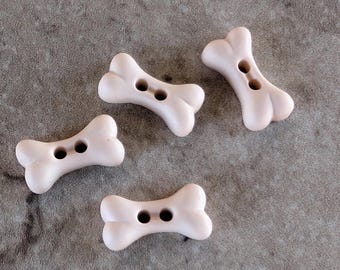 May include: Four light beige buttons shaped like dog bones. Each button features a smooth, matte finish and two circular holes positioned in the center for attachment. The buttons are arranged in a scattered pattern, showcasing their distinct shape.