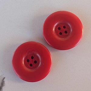 May include: Two round, bright red buttons with four holes each. The buttons have a slightly textured surface and a classic design. These buttons are suitable for sewing projects and crafts.
