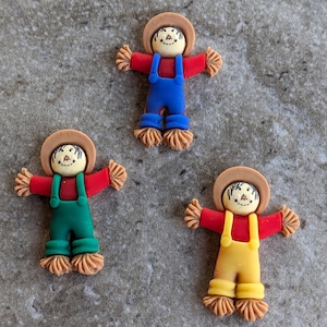 May include: Three small, handmade scarecrow figurines. The scarecrows are wearing different colored overalls: blue, green, and yellow. Each scarecrow has a brown hat and a painted face.