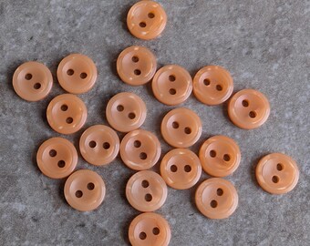May include: A collection of twenty-one round, peach-colored plastic buttons, each featuring two central holes and a slightly raised outer rim. The buttons are arranged in a loose cluster, displaying a smooth, semi-translucent finish throughout.