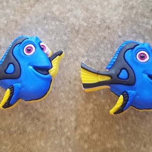 May include: Two blue and black cartoon fish with yellow fins and pink eyes. The fish are smiling and have a 3D appearance.