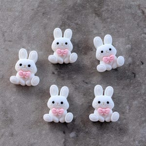 May include: Five small, white, rabbit-shaped embellishments, each featuring a soft pink bow tie at the neck, black eyes, and a small pink nose. The rabbits are depicted in a seated position with upright ears, designed as flat-backed decorative pieces.