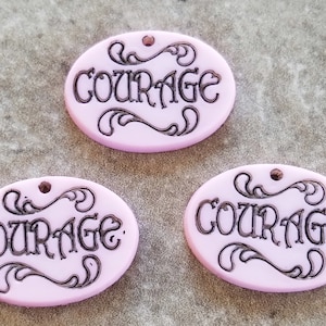 May include: Three pink oval-shaped charms with the word "COURAGE" written in black script. Each charm has a small hole at the top for hanging.