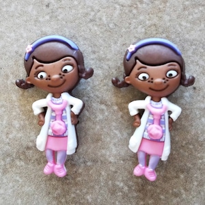 May include: Two brown-skinned cartoon doctor dolls with purple headbands and pink dresses. The dolls are wearing white lab coats and have a stethoscope around their necks.
