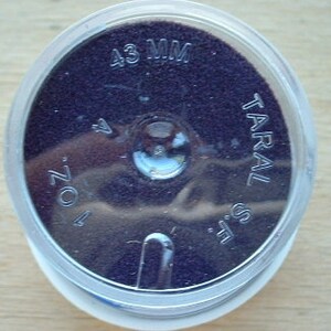 May include: A close-up of a small, round, clear plastic container with a purple, textured surface inside. The container is labeled "43 MM" and "TARAL S.L."