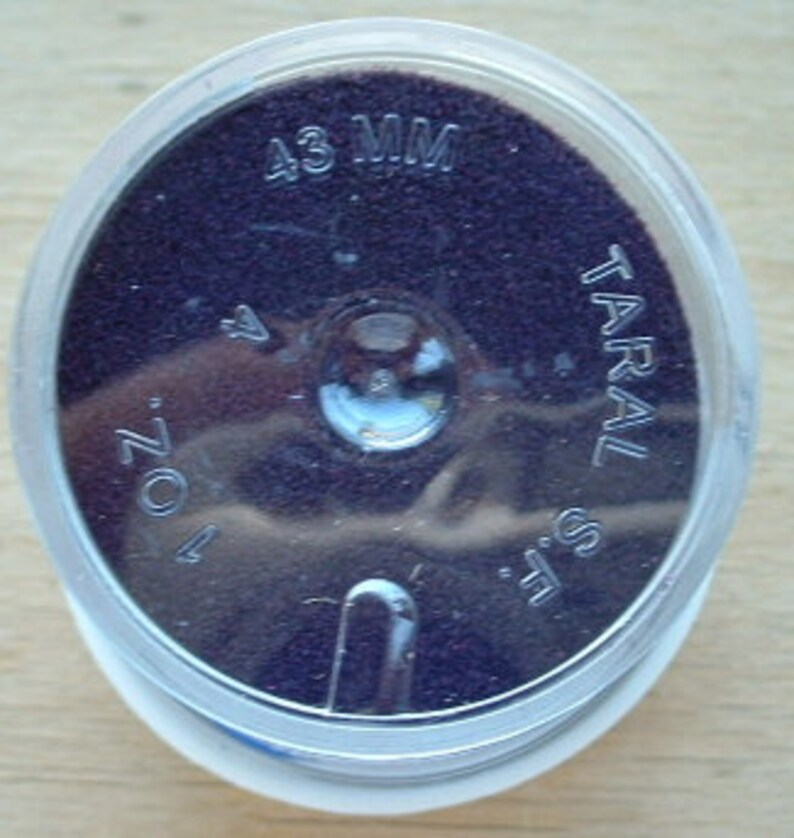 May include: A close-up of a small, round, clear plastic container with a purple, textured surface inside. The container is labeled "43 MM" and "TARAL S.L."