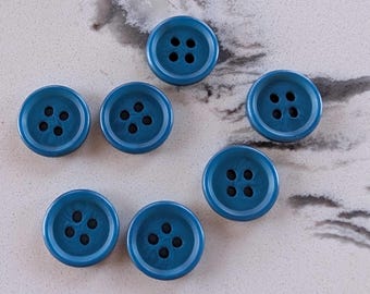 May include: Seven teal-colored buttons with four holes each are scattered on a light-colored surface. The buttons have a glossy finish and a slightly textured appearance. The buttons are round and appear to be made of plastic.