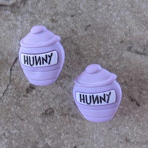 May include: Two light purple, cartoon-style honey pots with lids. Each pot has a white label with the word "HUNNY" in black letters. The pots are on a textured, gray surface.