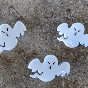 May include: Three white ceramic ghost figurines with different facial expressions. The ghosts are cut out and have a flat back.