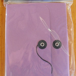 May include: A purple accordion keeper with a black string and two black buttons. The packaging says "Bazzill Basics Paper" and "bazzill accordion keeper".