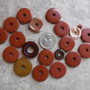 May include: A collection of 18 round, flat, red, brown, and tan stone beads with holes in the center. The beads are arranged in a circle on a gray surface. A silver US quarter is included for scale.