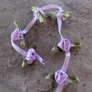 May include: Close-up of several lavender satin ribbon roses with green satin leaves. The roses are connected by lavender ribbon. The image is taken on a gray stone surface. These decorative flowers are ideal for crafts and embellishments.