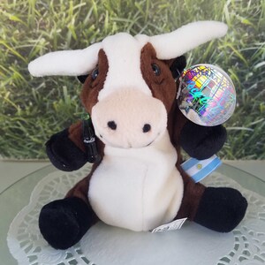 May include: Brown and white plush cow holding a Coca-Cola bottle and a globe with the text "Inter-America" on it. The cow is sitting on a white doily.