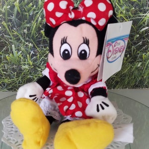 Może przedstawiać: Pluszowa zabawka Minnie Mouse w czerwonej sukience w białe kropki i czerwoną kokardkę w białe kropki. Zabawka ma ż&oacute;łte buty i białą metkę z napisem "Disney Store".