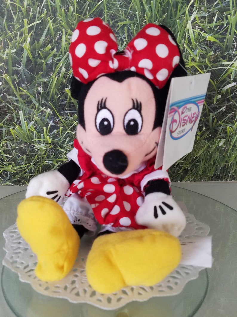 Może przedstawiać: Pluszowa zabawka Minnie Mouse w czerwonej sukience w białe kropki i czerwoną kokardkę w białe kropki. Zabawka ma ż&oacute;łte buty i białą metkę z napisem "Disney Store".