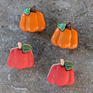 May include: Four ceramic pumpkin ornaments in orange and red with green stems. The pumpkins are arranged on a gray textured surface.