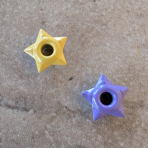 May include: Two star-shaped candle holders. One is yellow, the other is purple. Each has a central hole for a candle. The candle holders are on a textured, neutral-toned surface.