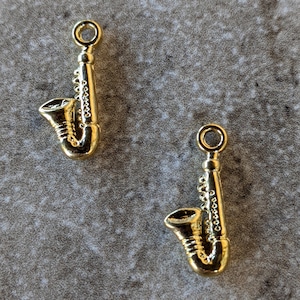 May include: Two gold-colored saxophone charms. The charms are detailed with a saxophone shape and have a loop at the top for attaching to a necklace or bracelet.