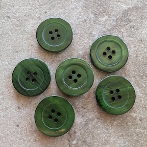 May include: A set of six round, dark olive green buttons, each featuring a smooth, slightly glossy finish and four central holes for attachment. The buttons exhibit a subtle, linear grain pattern across their circular surfaces.