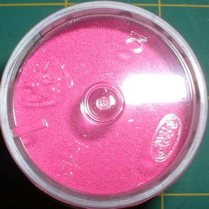 May include: A small, round container filled with bright pink sand. The container has a clear lid with a small hole in the center.